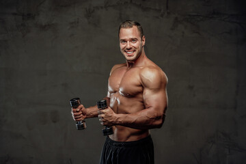 Fototapeta na wymiar Brutal handsome bodybuilder with two dumbells staying and smiling in special grung background.