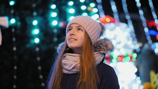 Teenage girl dressed in warm clothes looks up at the Christmas lights in the city. Happy holidays