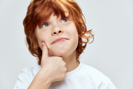 Red-haired boy holds his hand on his face and looks up smile white t-shirt close-up 