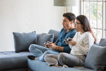Obraz premium Handsome Caucasian man and beautiful Asian woman lover sitting in house living room and using joystick to play game togehter with happiness and joyful. Diverse couple and stay at home concept