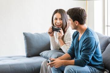 Fototapeta premium Handsome Caucasian man and beautiful Asian woman lover sitting in house living room and using joystick to play game togehter with happiness and joyful. Diverse couple and stay at home concept