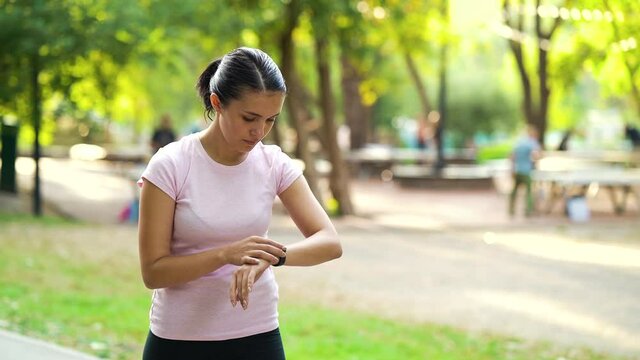 Fit woman in sportswear looking at fitness bracelet and starting running in summer park, blurred people on background. Active female checking heart rate before training. Concept of sport