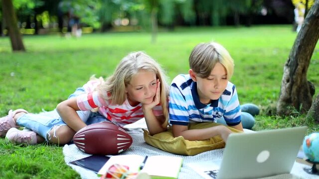 Bored children looking at laptop screen resting outdoor in city park on beautiful summer sunny day. Online education concept. Technology for kids. Boy and girl using notebook on vacations. Traveling 