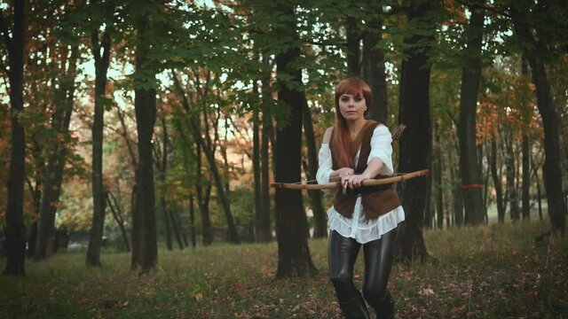 A woman archer hides near a tree and aims an arrow from a bow. Medieval girl hunter in a dense green forest. Сostume of a vintage warrior with a weapon in his hands. Fantasy shooting, video footage