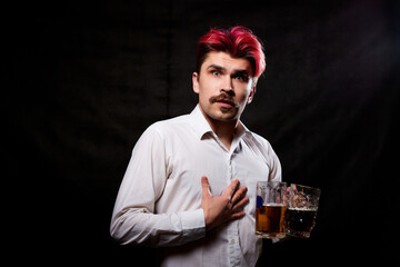 Young handsome guy with red hair in white shirt drinking beer. Funny man with emotions on the face and beer mug in hand and black background. Alcoholic is happy with alcohol