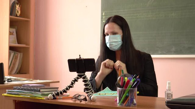 A young teacher leads a lesson, filming himself on a smartphone. Female teacher wearing protective face mask.Video tutorial during quarantine.Covid-19.Finger counting.4k