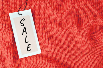 Word Sale on price tag on orange knitted wool fabric with folds or warm sweater. A light cardboard label card attached to the black rope on the clothes. Shopping concept, background with copy space.