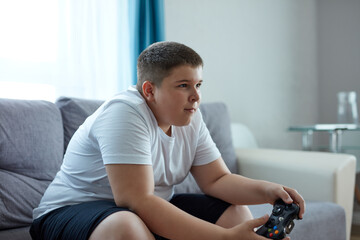 keen caucasian boy playing video games, overweight boy sit on sofa alone, has problems with nutrition, lead unhealthy lifestyle. laziness concept
