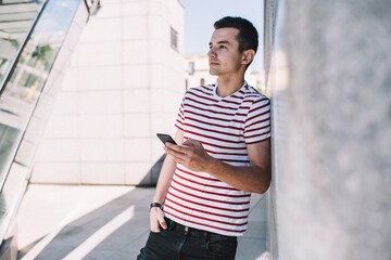 Optimistic male freelancer surfing phone and leaning on stone fence in urban street