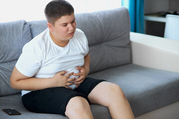 fat teenager boy has stomach ache after junk food, sit on sofa holding belly, suffer from pain