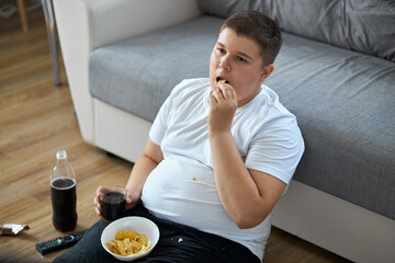 overweight fat boy eat junk food while watching tv alone at home, sit on the floor with french...