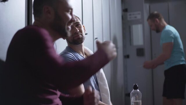 Positive Caucasian sportsmen sitting in locker room after training and talking. Smiling handsome athletic men chatting and gesturing as resting after workout. Masculinity concept.