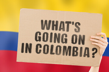 The question " What's going on in Colombia? " on a banner in men's hand with blurred Colombian flag on the background. Protests on the street. Riot. Violence. Economic crisis. Collapse. Politics