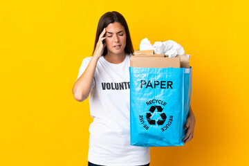 Young brazilian girl holding a recycling bag full of paper to recycle isolated on yellow background with headache