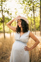 Pregnant woman posing outdoors in the trees with sunset light with hat.