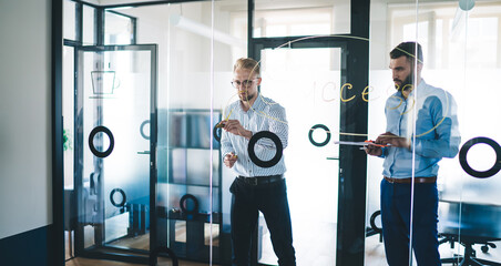 Professional male entrepreneurs writing notes collaborating on finance and business during meeting, young Caucasian men colleagues explaining ideas and opinions writing on modern glass board