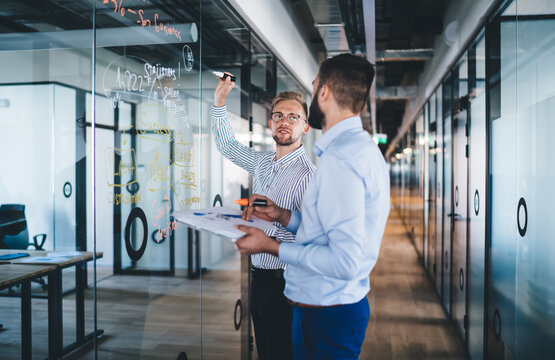 Confident male boss looking at his employee explaining data and information writing on glass and communicating, young 20s men business partners solving problem having productive job 