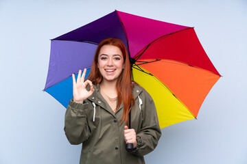 Redhead teenager girl holding an umbrella over isolated blue background showing an ok sign with fingers