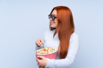 Redhead teenager girl over isolated blue background with 3d glasses and holding a big bucket of popcorns