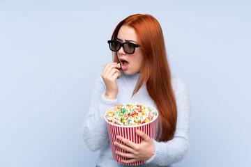 Redhead teenager girl over isolated blue background with 3d glasses and holding a big bucket of popcorns while looking side