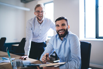 Portrait of cheerful male employees in formal outfit sitting at table desktop with laptop collaborating together for analyzing accounting for startup, excited professional men laughing at camera
