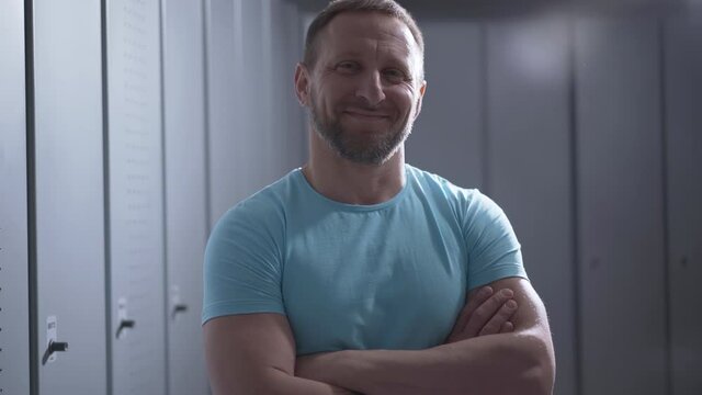 Portrait of confident sportsman crossing hands and smiling at camera. Adult bearded Caucasian man posing in gym locker room. Positive guy standing in dressing room. Sport concept.