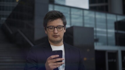 Man holding the phone on background of corporate business centre buildings. Man dressed in black suit and white shirt scrolling the phone, middle shot, business university