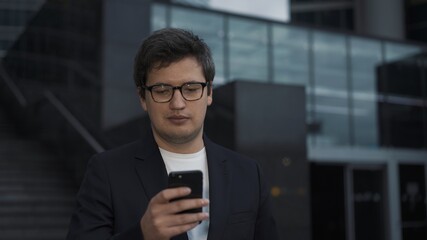 Man with a phone on background of corporate business centre buildings. Man dressed in black suit and white shirt scrolling the phone, middle shot, business university