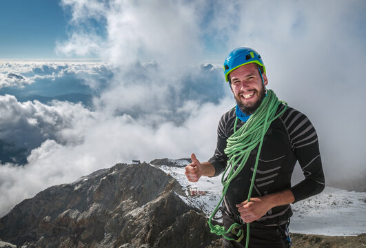 Smiling Climber in a safety harness, helmet, and  on body wrapped climbing rope with picturesque clouds background showing thumb sign at 3600m altitude during Mont Blanc ascending, France route