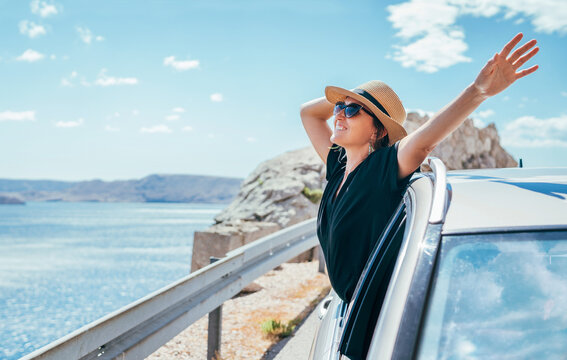 Cheerful Woman portrait enjoying the seaside road trip. Dressed a black dress, straw hat and sungllasses she wide opened arms and shining with happiness. Summer vacation traveling by auto concept.