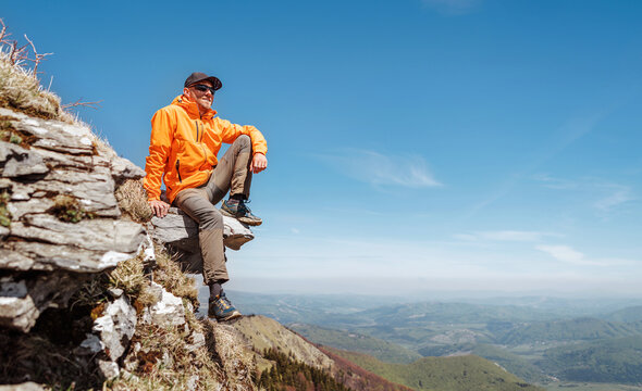 Dressed bright orange jacket hiker in baseball cap and sunglasses sitting on rocky cliff enjoying green valley at Mala Fatra mountain range,Slovakia. Active people and European tourism concept image.