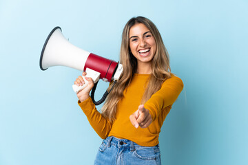 Young hispanic woman over isolated blue background holding a megaphone and smiling while pointing...
