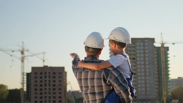 Father and son happy Family concept. Little boy and his dad engineer builder architect with safety helmets, on background new buildings and construction cranes on site. Future profession, 4 K slow-mo