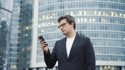 Young businessman looking at the phone on background of business centre buildings. Man dressed in black suit and white shirt scrolling the phone near corporate glass buildings