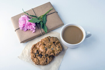 Romantic vintage still life with pretty gift box wrapped with brown craft paper and decorated with pink flower on white background. Dessert (oat cookies) served for tea or coffee break.