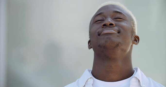 Portrait of happy African American man in white closes eyes, breathes in fresh air through nose, makes calm exhalation, practices breathing exercises, feels peaceful, shakes head to sides with relief