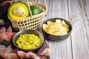 Guacamole with corn tortilla chips in plate and avocados in basket on wooden table