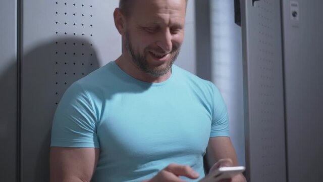 Smiling bearded sportsman using smartphone in gym locker room. Portrait of happy positive Caucasian man surfing Internet or using social media after workout. Lifestyle concept.