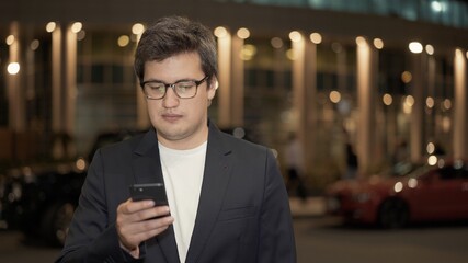 Businessman in eyeglasses texting on the phone on background of building with yellow lights, night shot man standing near the road. Middle shot of man in black jacket with a phone