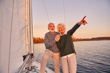 Happy senior couple holding hands and smiling while standing on the side of yacht deck floating in sea, woman pointing at the horizon, sailing at sunset