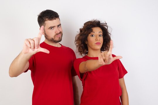 Young beautiful couple wearing red t-shirt on white background making fun of people with fingers on forehead doing loser gesture mocking and insulting.