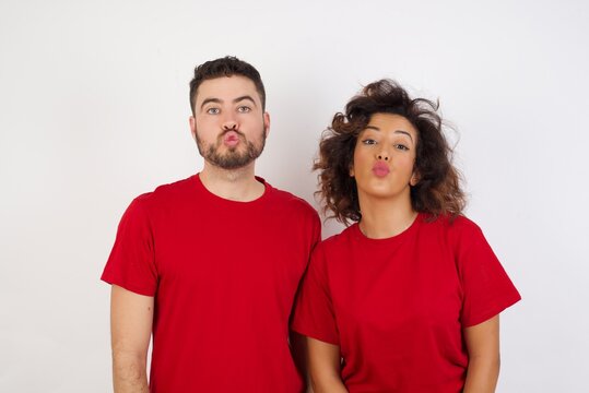 Shot of pleasant looking Young beautiful couple wearing red t-shirt on white background pouts lips, looks with green eyes at camera,  has fun with girlfriend, Human facial expressions