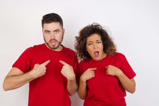 Photo of embarrassed Young beautiful couple wearing red t-shirt on whi indicates at theself with puzzled expression, being shocked to be chosen to participate in competition, hesitates about something