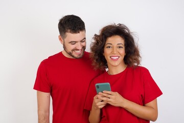 Young beautiful couple wearing red t enjoys distant communication, uses mobile phone, surfs fast unlimited internet, has pleasant smile, makes shopping online,