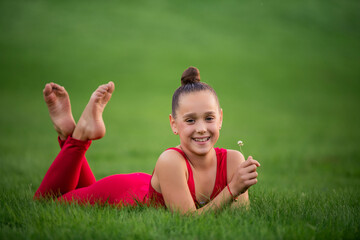 girl schoolgirl gymnast in bright red overalls is resting on grass in the park, holding a clover flower in her hands, laughing. She has braces on her teeth.