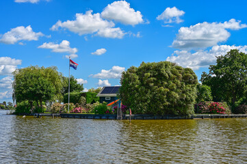 Island with a house and the Dutch flag on it in a lake with green water. Above it a bright blue sky with small white clouds. Reeuwijk, The Netherlands, Europe.