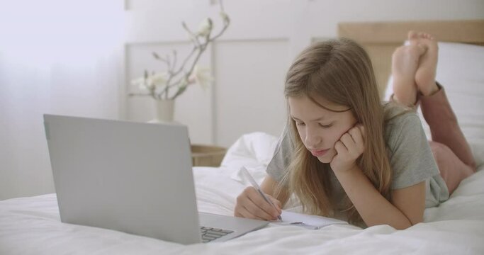 little girl is drawing and writing in copybook, lying on bed in front of laptop in her room, waiting online lesson