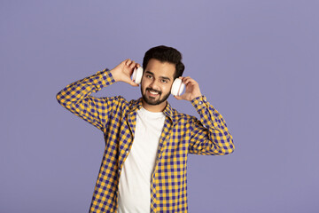 Obraz premium Smiling Indian guy listening to his favorite music in headphones on lilac background