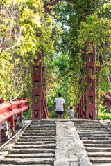 Bottom view of gray brick stone stair steps leading to red iron suspension pedestrian bridge in early autumn forest wood park. Natural landscape concept. People man boy far away in the background