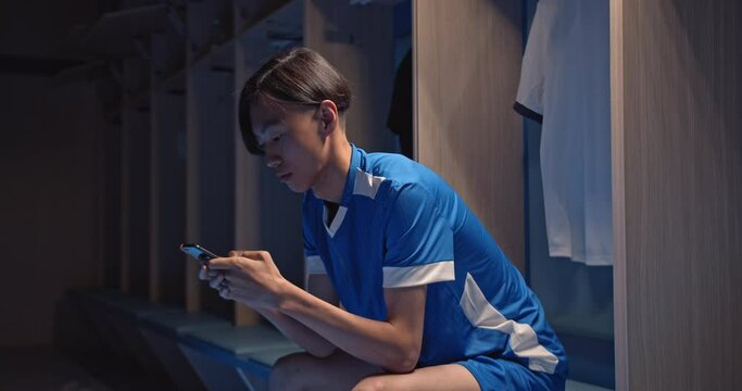 Asian Football fan holding phone and betting in internet with the smart phone at empty fitting room, soccer player, man playing games on phone after match, professional game, online bets and casino